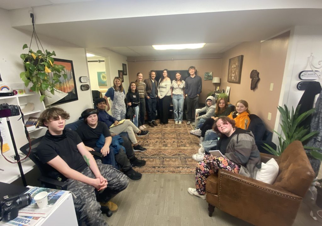 Group of young adults gathered in a cozy living room, some seated on chairs and others standing, smiling at the camera.