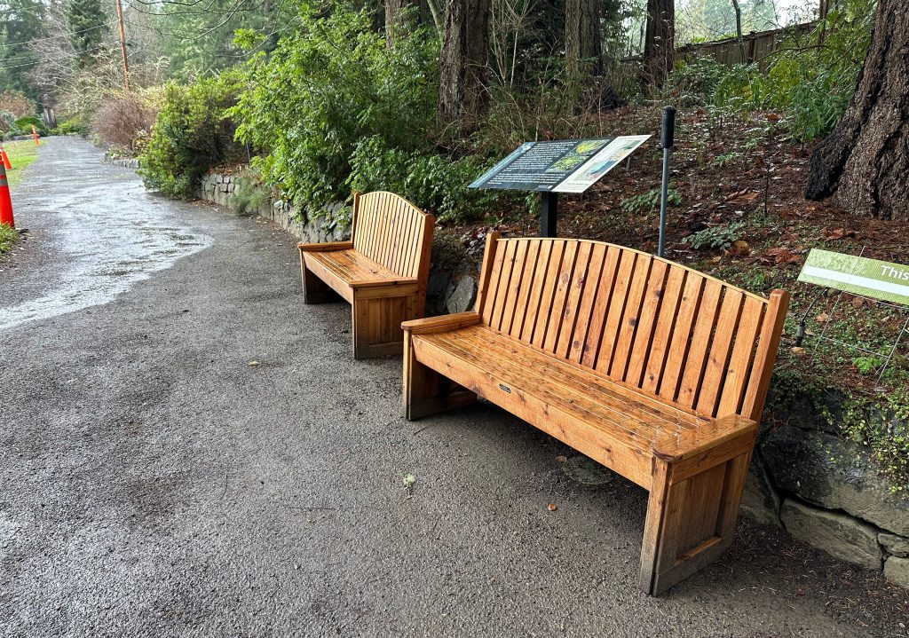 Two wooden benches with vertical slats sit along a paved path surrounded by greenery and informational signs in a park setting.