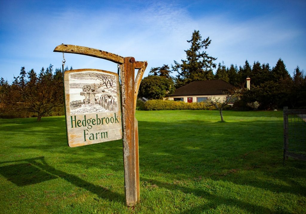 Rustic wooden sign reading 'Hedgebrook Farm' stands on a lush green lawn with a farmhouse and trees in the background under a clear blue sky.