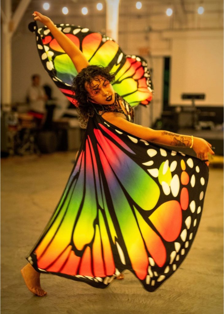 Person dancing barefoot indoors wearing vibrant butterfly wings with rainbow colors and black-and-white patterns.