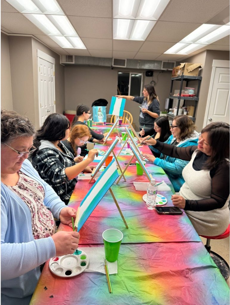Group of people seated at a long table painting on canvases in a well-lit room during an art class.