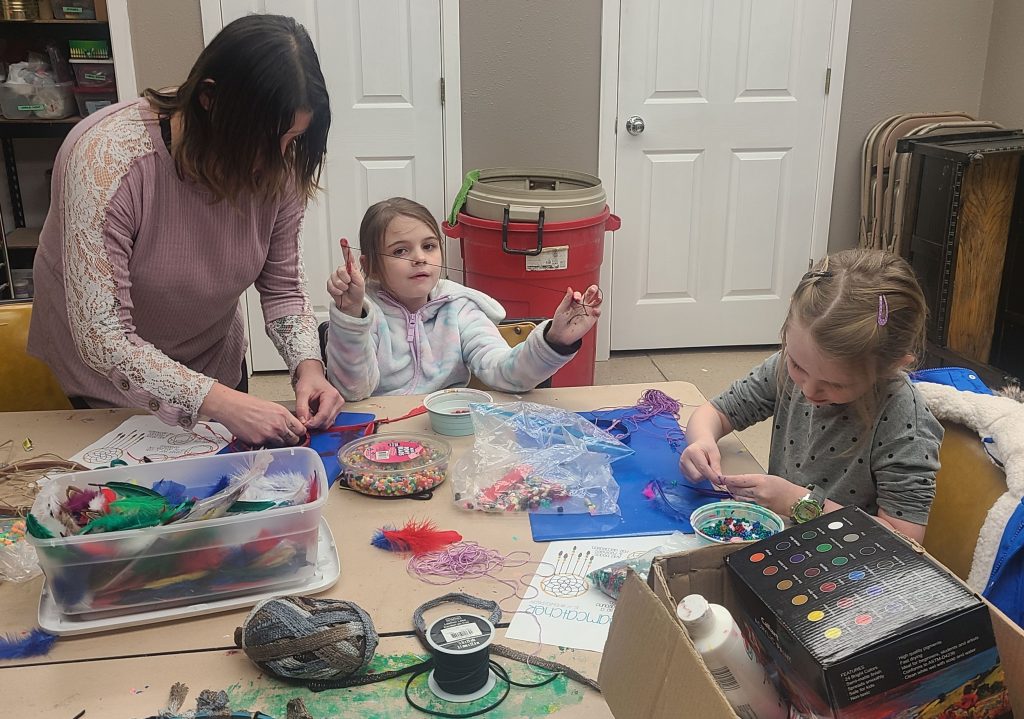 Two children and an adult crafting with beads, feathers, and colorful materials at a table in a cozy room.