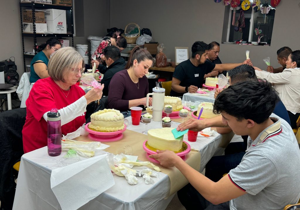 Group of people decorating cakes with frosting and piping bags around a table in a workshop setting.
