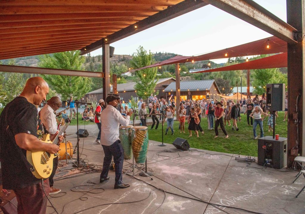 Live band performing under a pavilion with a crowd dancing and enjoying music on a grassy area at sunset.