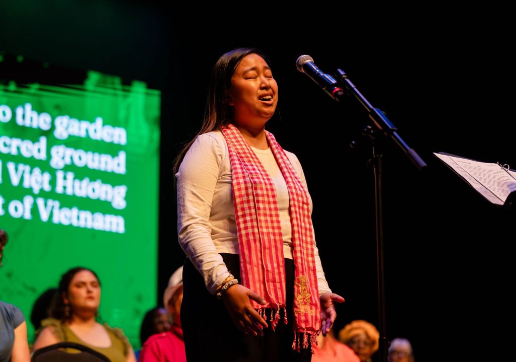 Woman wearing a red checkered scarf stands at a microphone on stage during a performance or speech event.