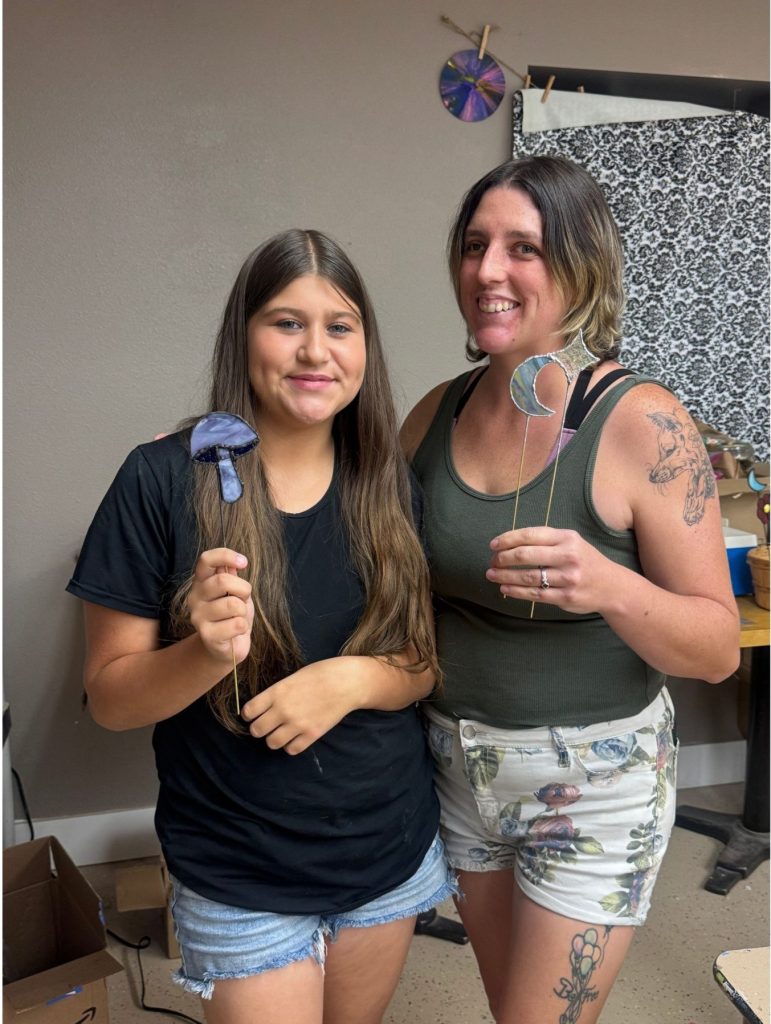 Two women standing indoors, each holding a colorful lollipop, one wearing a black shirt and denim shorts, the other in a green tank top and patterned shorts with tattoos.