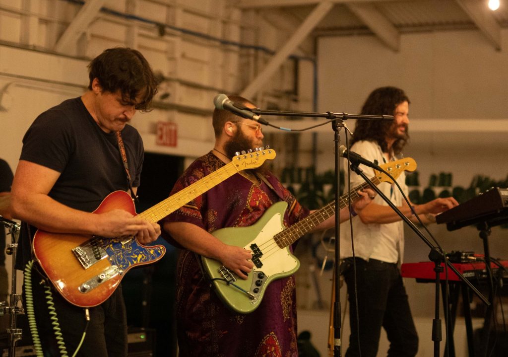 Three musicians performing indoors with electric guitar, bass guitar, and keyboard under warm lighting.