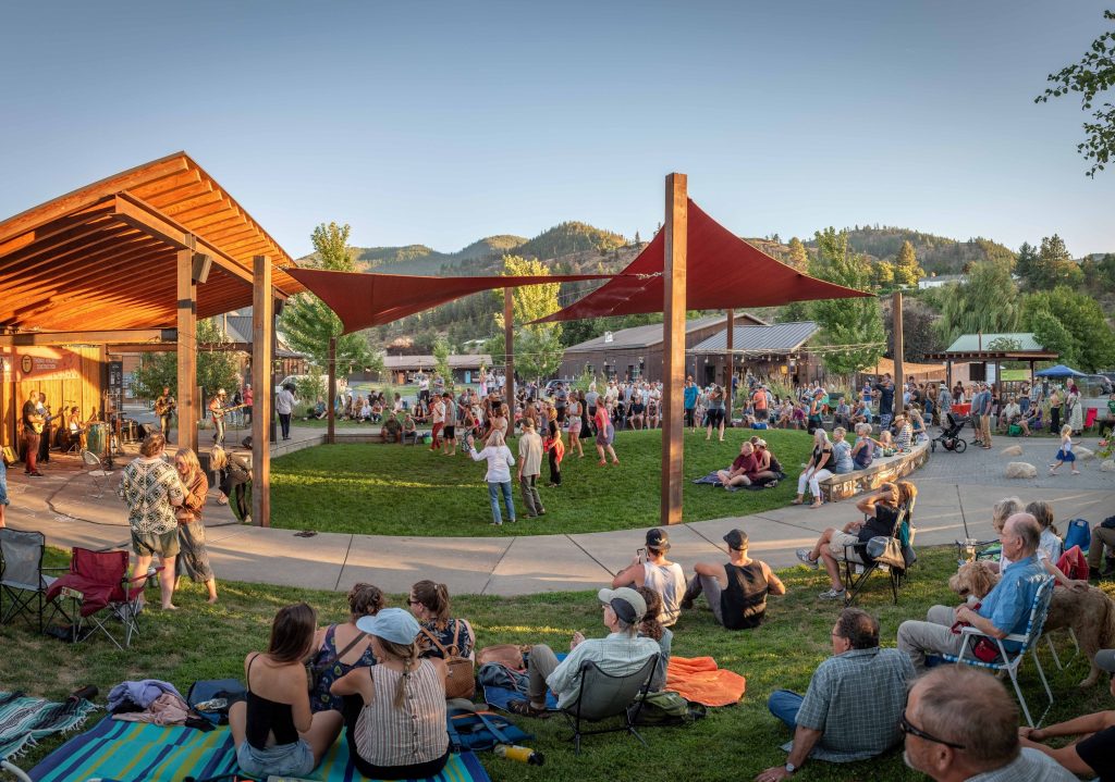 Crowd enjoying an outdoor event on a grassy area with red shade sails and mountains in the background during clear weather.