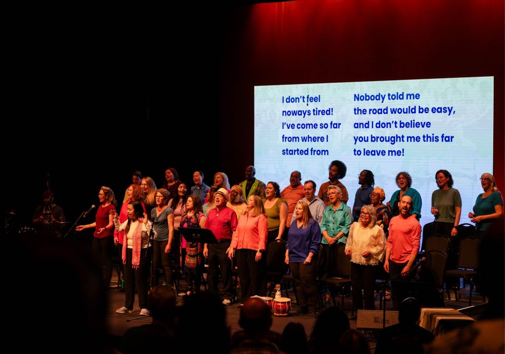 Tacoma Refugee Choir performs on stage under red lighting with song lyrics projected behind them.