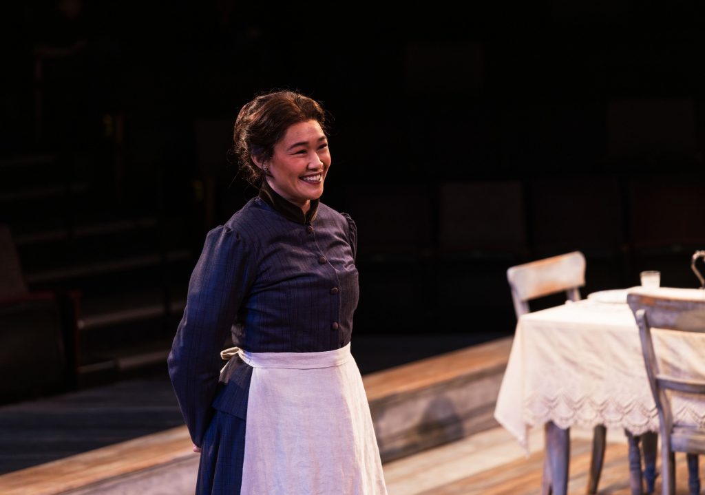 Woman in a dark blue dress and white apron stands on a wooden stage near a table with a lace tablecloth and chairs.