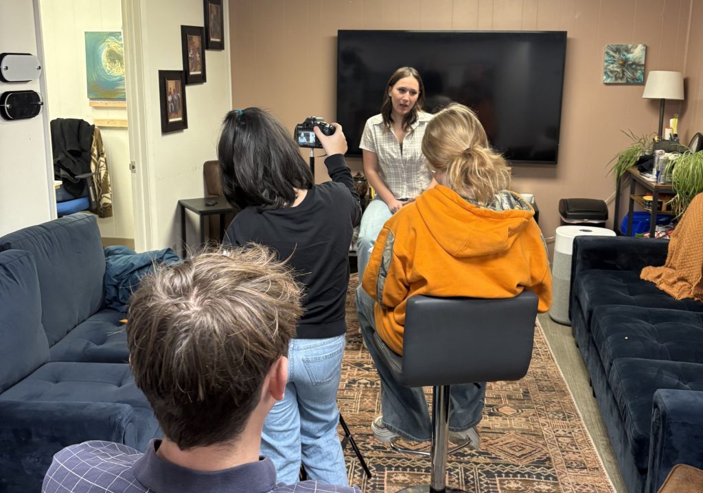 Person sitting on a stool facing a camera held by another individual in a cozy living room setup with a large TV and blue couches.