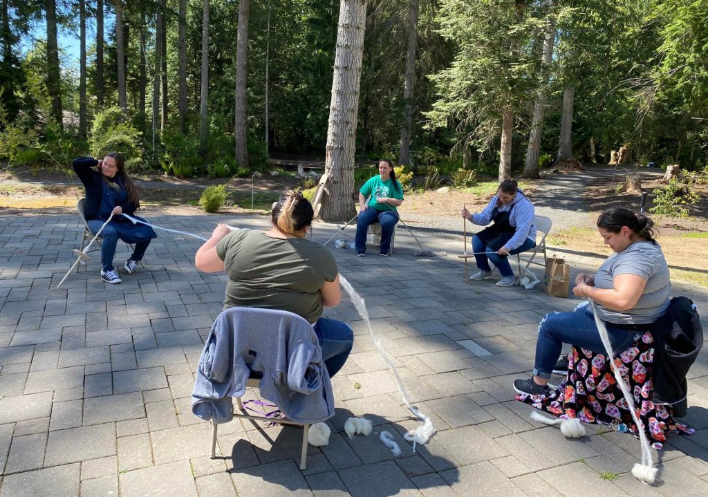 Five people sitting in a circle outdoors on folding chairs, engaged in a knitting or crochet activity on a paved area surrounded by trees.
