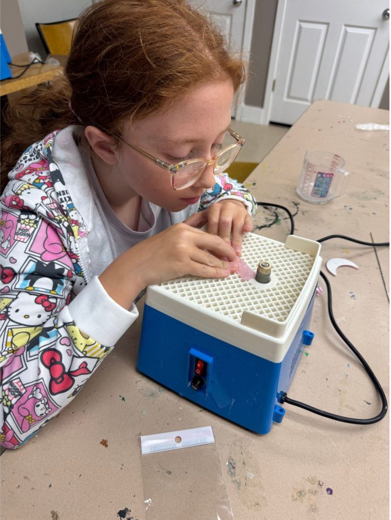 Child wearing a colorful Hello Kitty hoodie focused on using a blue and white pottery wheel at a craft table.