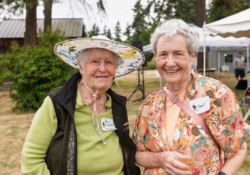 Two elderly women outdoors at a casual gathering, one wearing a wide-brimmed floral hat and green sweater, the other in a colorful floral blouse holding a drink.