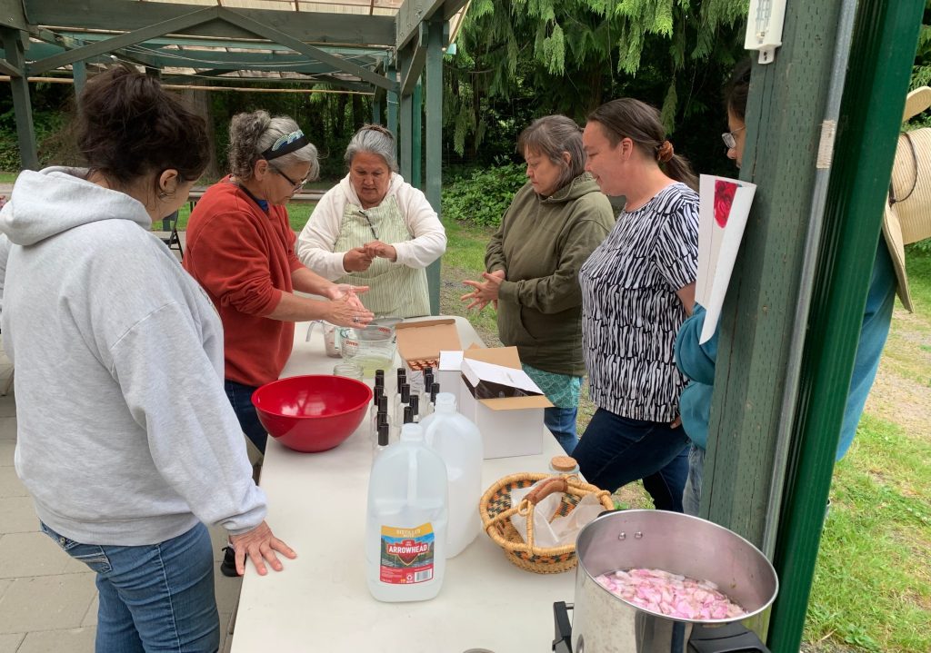 Group of five people gathered around a table outdoors, preparing ingredients with bottles and a large pot of pink petals nearby.