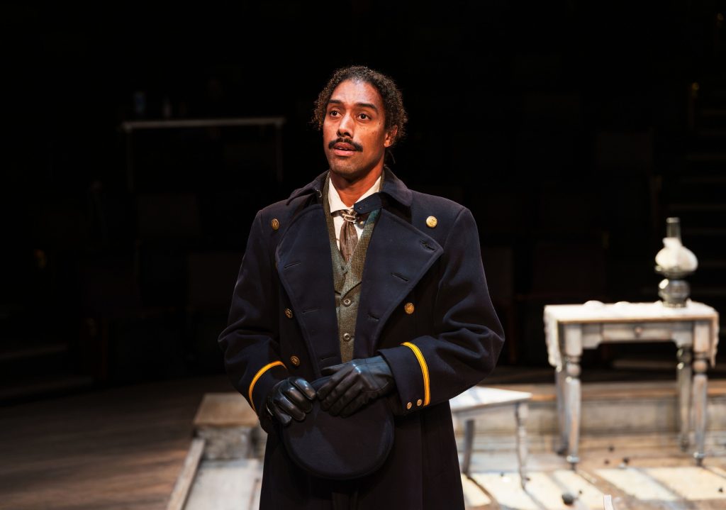 Actor in a dark navy double-breasted coat with gold buttons and gloves stands on a dimly lit stage set resembling a vintage room.