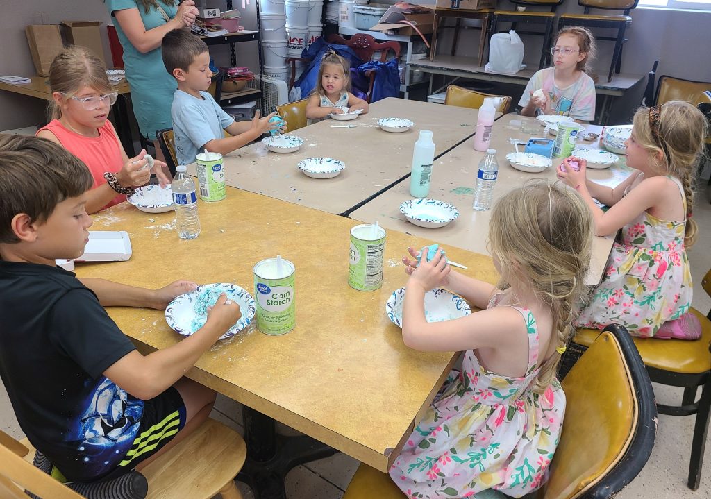 Children sitting around a table engaged in a hands-on activity with craft supplies and paper plates in a classroom setting.