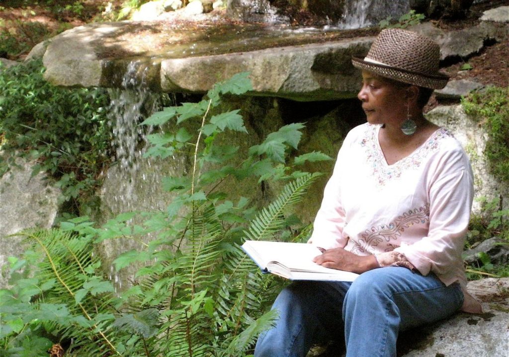 Woman wearing a straw hat and light pink blouse sitting on a rock near a small waterfall, writing in a notebook surrounded by green foliage.