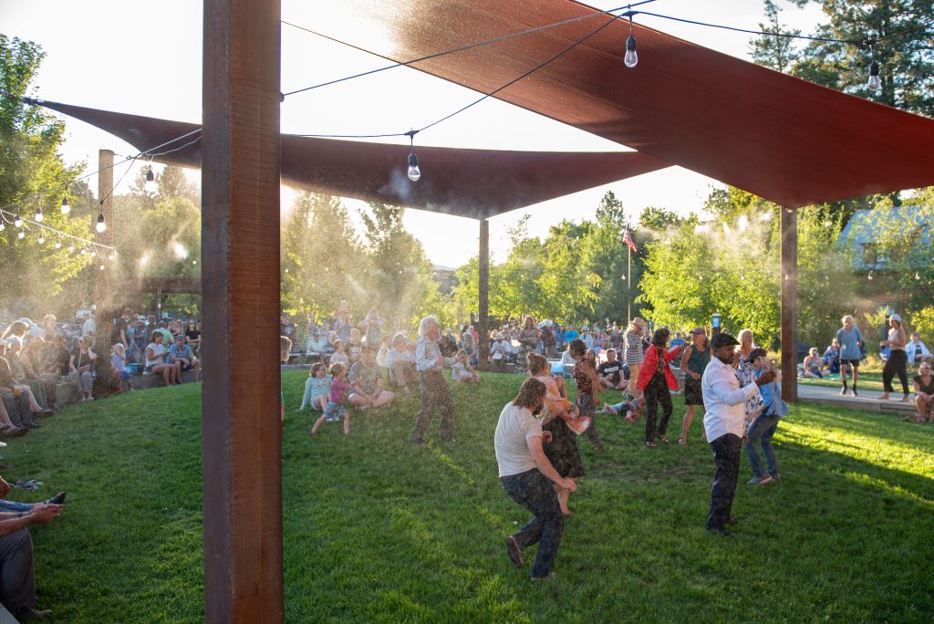 Families and children enjoying a sunny day dancing and playing on a grassy area under shade sails in a park.
