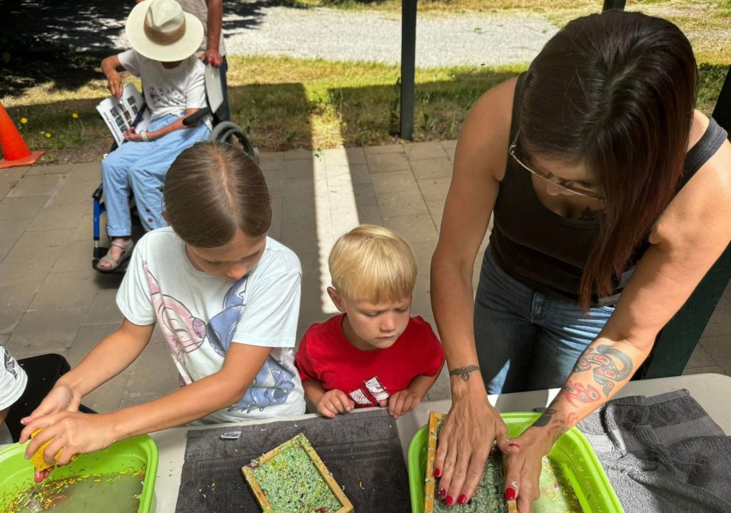 A woman and two children engaging in a hands-on activity with green trays and textured materials on a table outdoors.