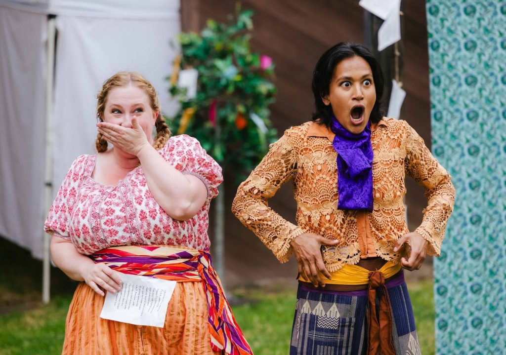 Two women dressed in colorful traditional attire stand outdoors, one holding a paper and the other adjusting her clothing.