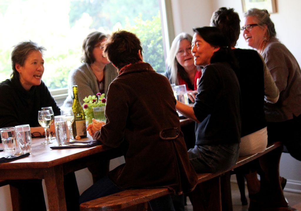 Group of six people sitting around a wooden table engaged in conversation with drinks and a bottle of wine present.