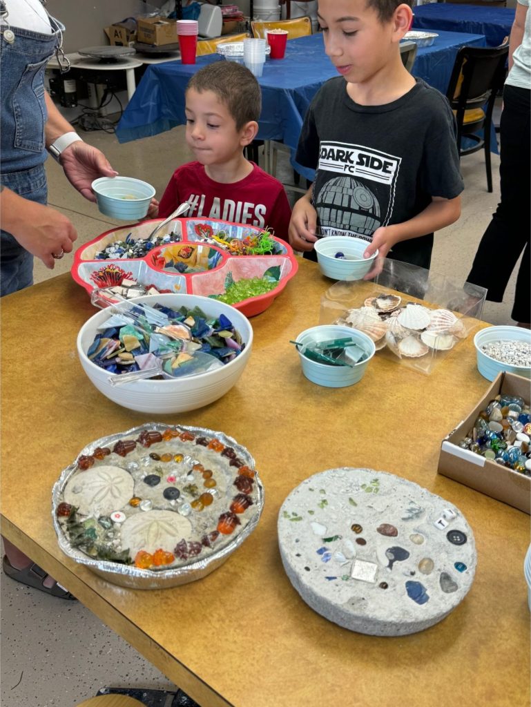 Two children stand at a table filled with bowls of colorful stones, shells, and craft materials for an art project.