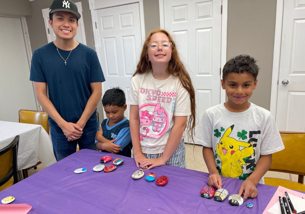 Four children standing and sitting around a purple-covered table displaying decorated computer mice in a home setting.