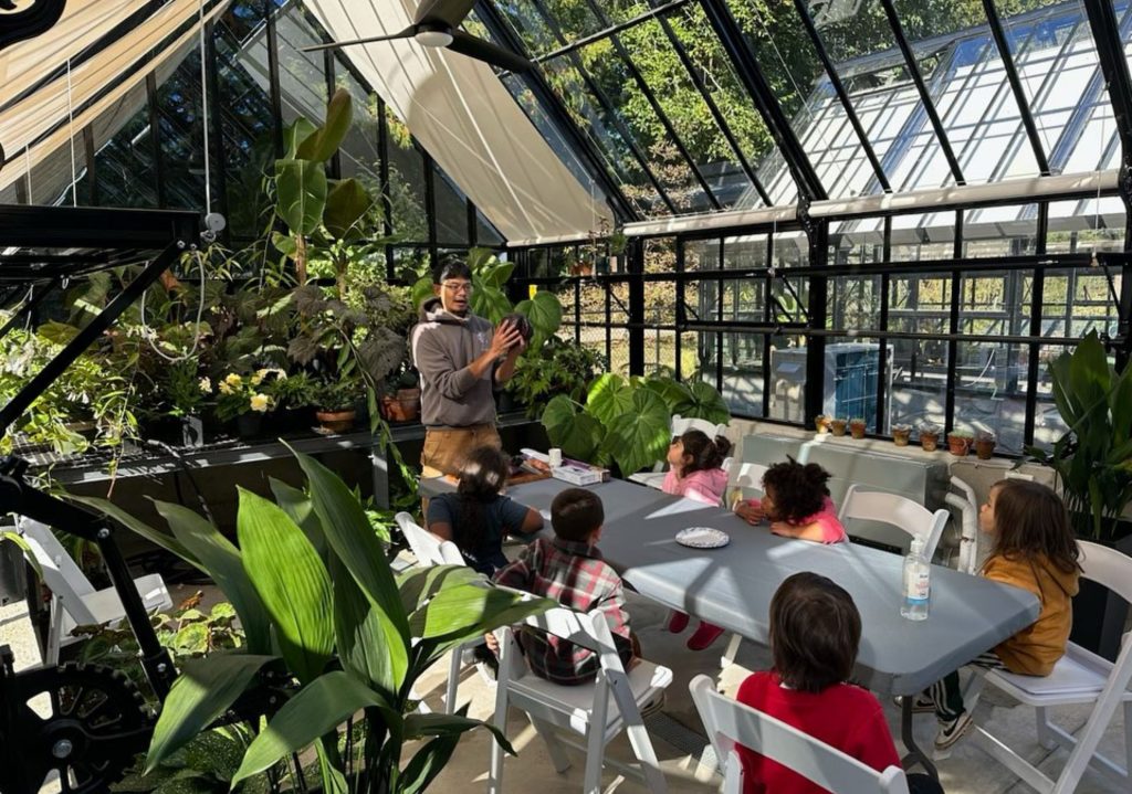 Teacher explaining plant biology to children seated around a table inside a sunlit greenhouse filled with lush greenery.