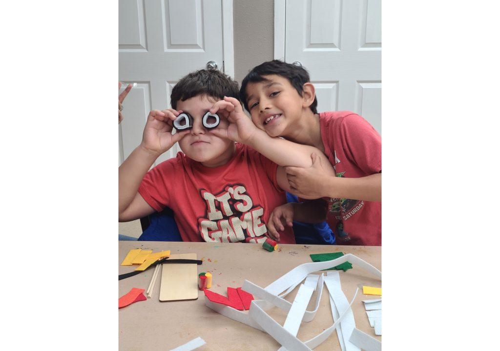 Two children in red shirts sitting at a table with paper craft materials and colorful cutouts scattered around them.