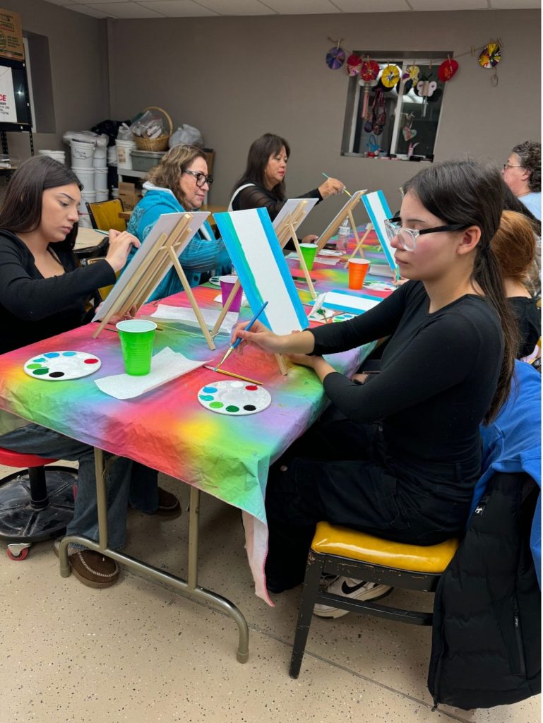 Group of women seated around a table covered with a colorful cloth, painting on canvases during an art class.