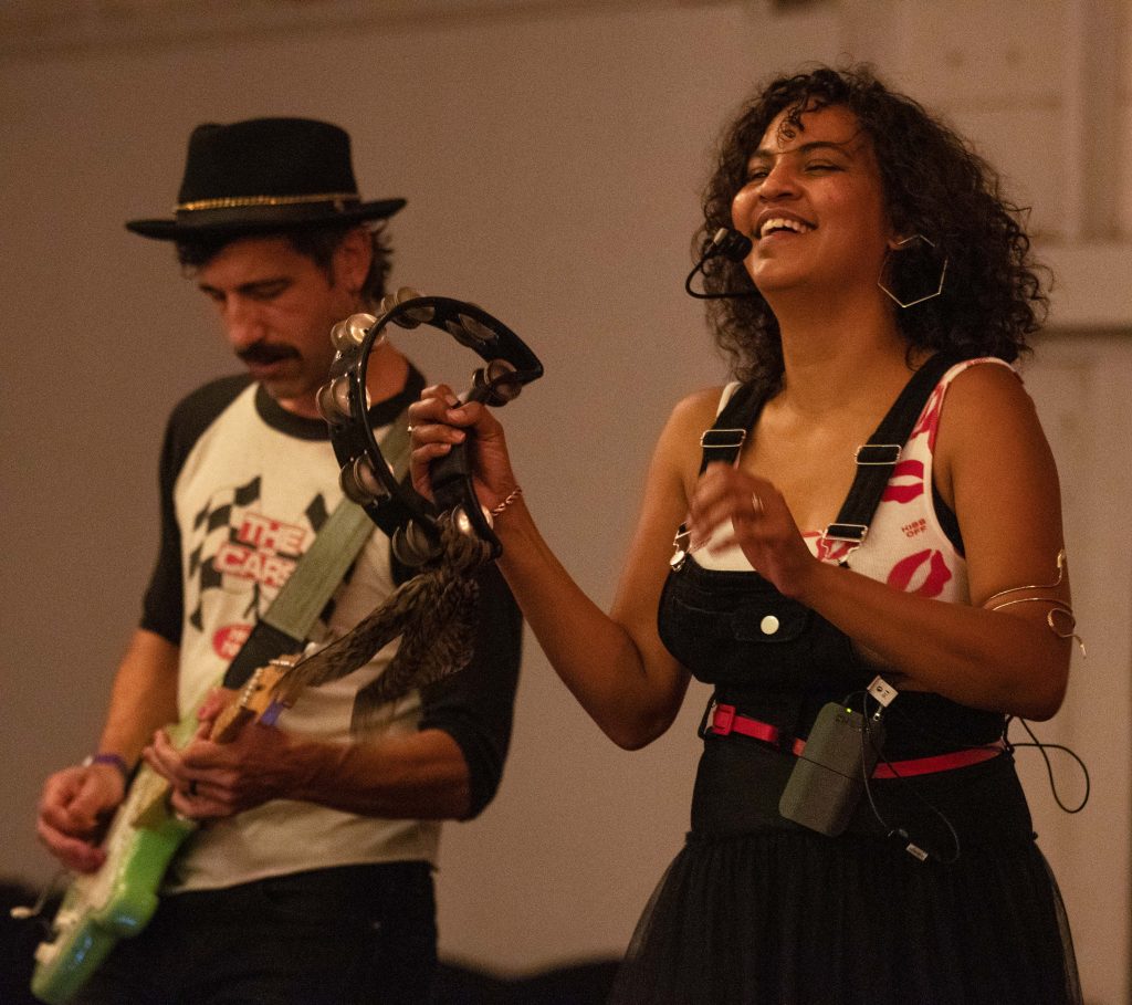 Female singer playing tambourine and male guitarist performing on stage in casual attire.