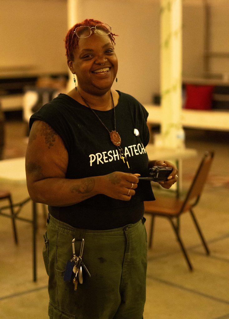 Person wearing a sleeveless black shirt with 'PRESH:ATECH' text, holding a camera in an indoor setting with chairs and tables.