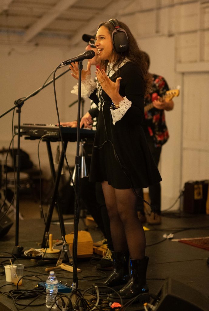 Female singer in a black dress with lace cuffs performing on stage wearing headphones and gesturing expressively into a microphone.