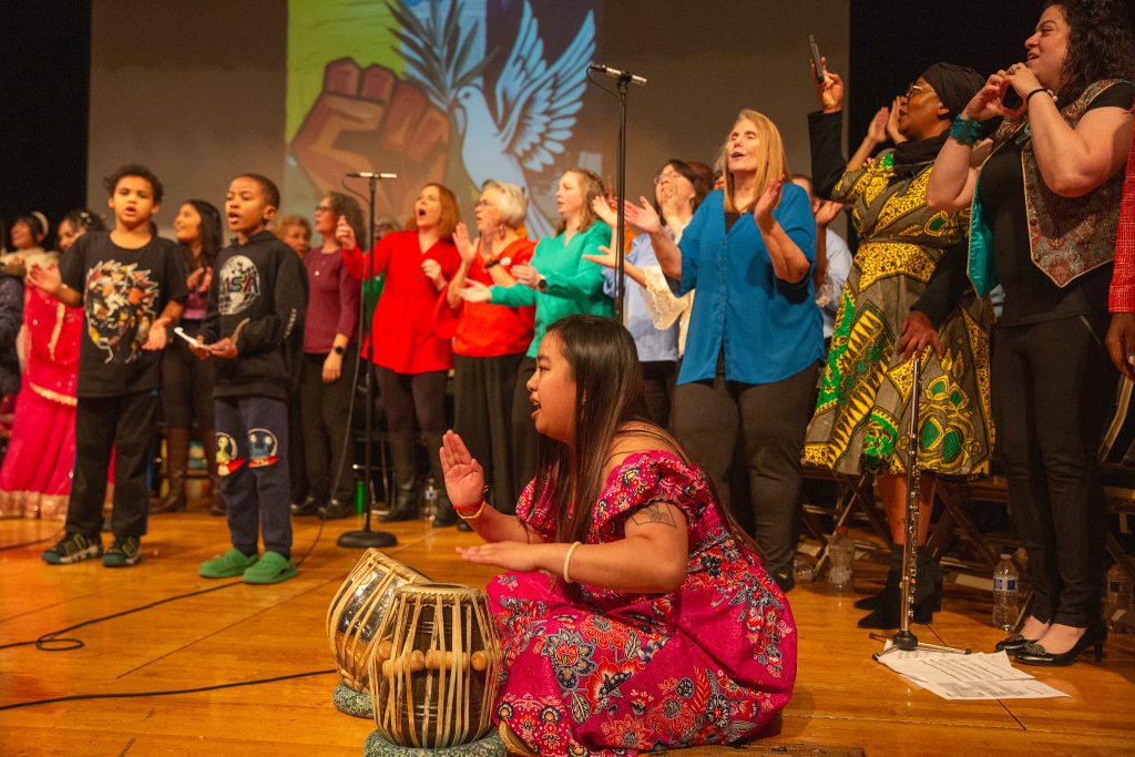 The Tacoma Refugee Choir sing on stage with a woman in colorful attire playing a traditional drum in front.