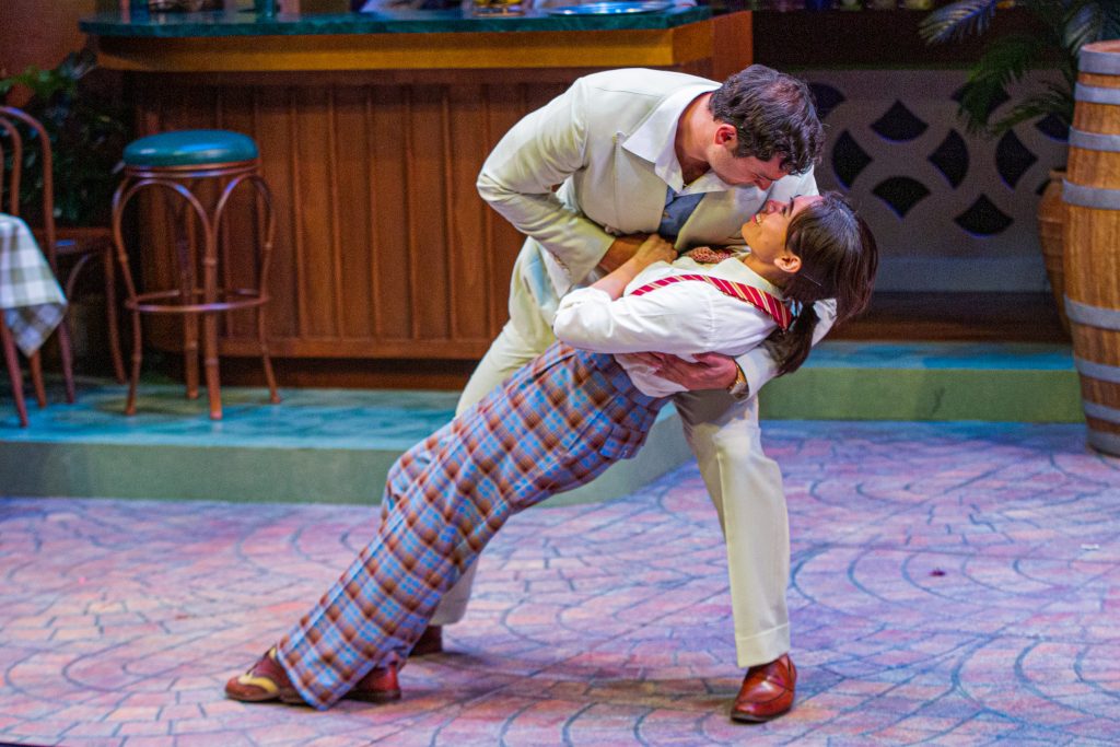 A man in a white suit dips a woman in plaid pants and suspenders as they share a romantic gaze on stage.