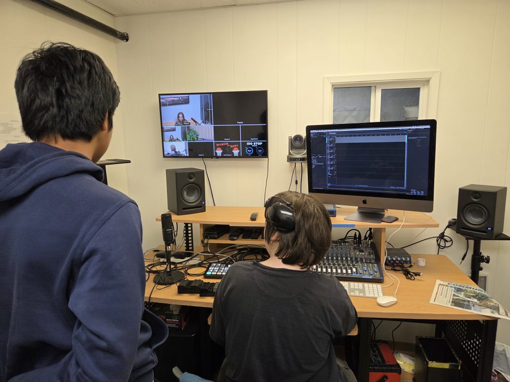 Two people in a recording studio, one seated at a mixing console with headphones, the other standing nearby watching a video call on a wall-mounted screen.
