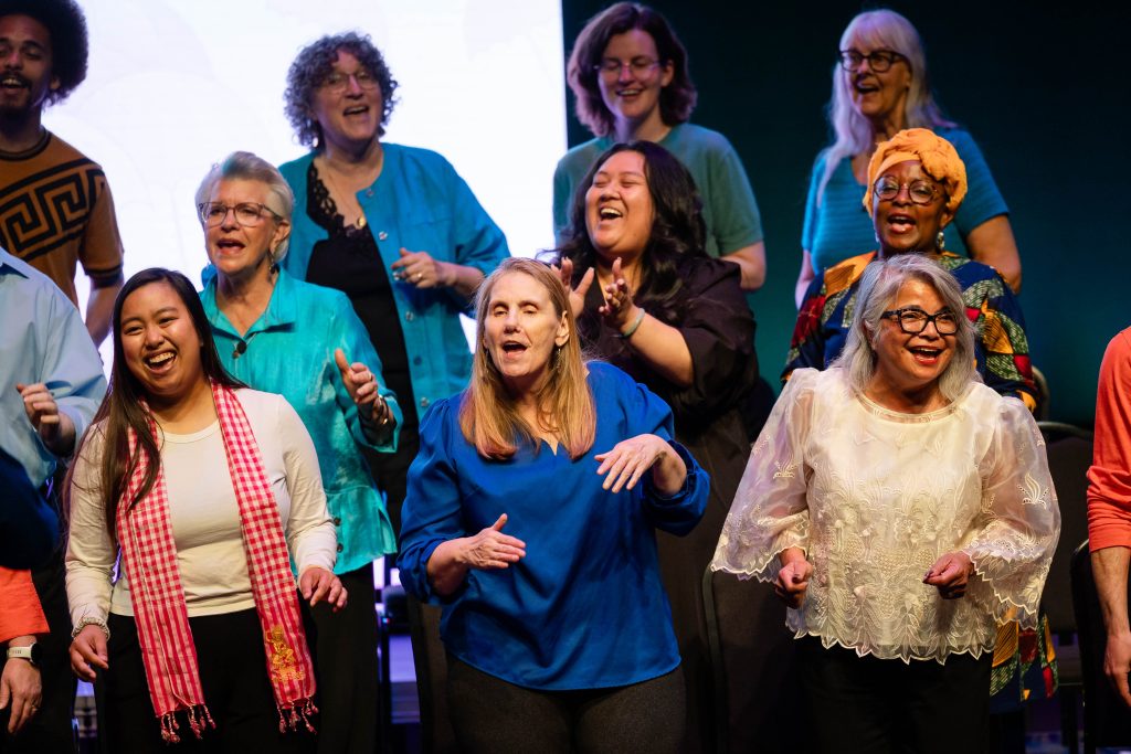 Women from the Tacoma Refugee Choir women stand on stage, engaged in a lively discussion or performance under bright lighting.