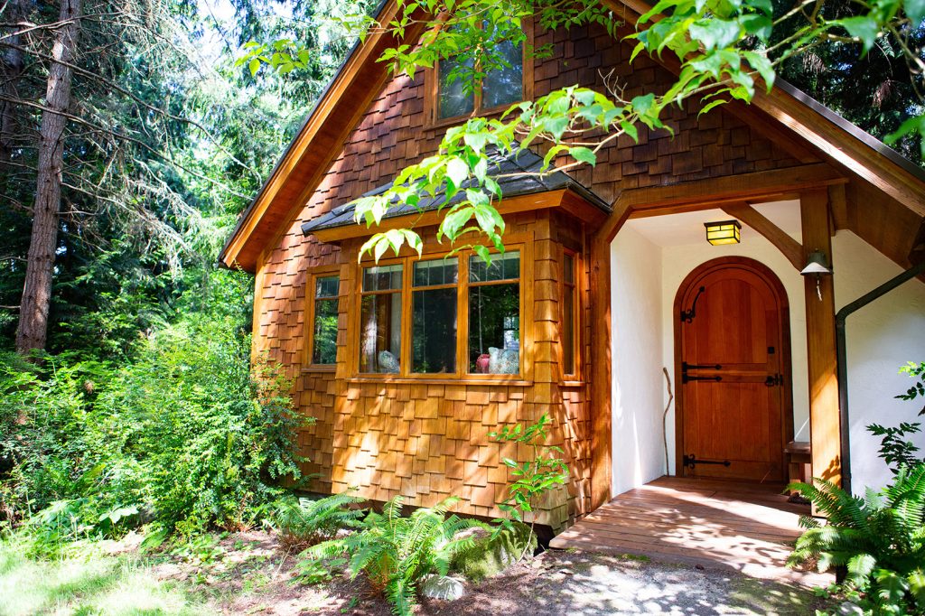 Cozy wooden cottage nestled in a lush green forest with sunlight filtering through the trees onto the front porch.