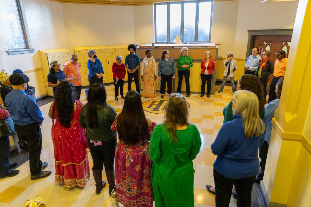 The Tacoma Refugee Choir stands in a large circle inside a bright room with yellow walls and windows.