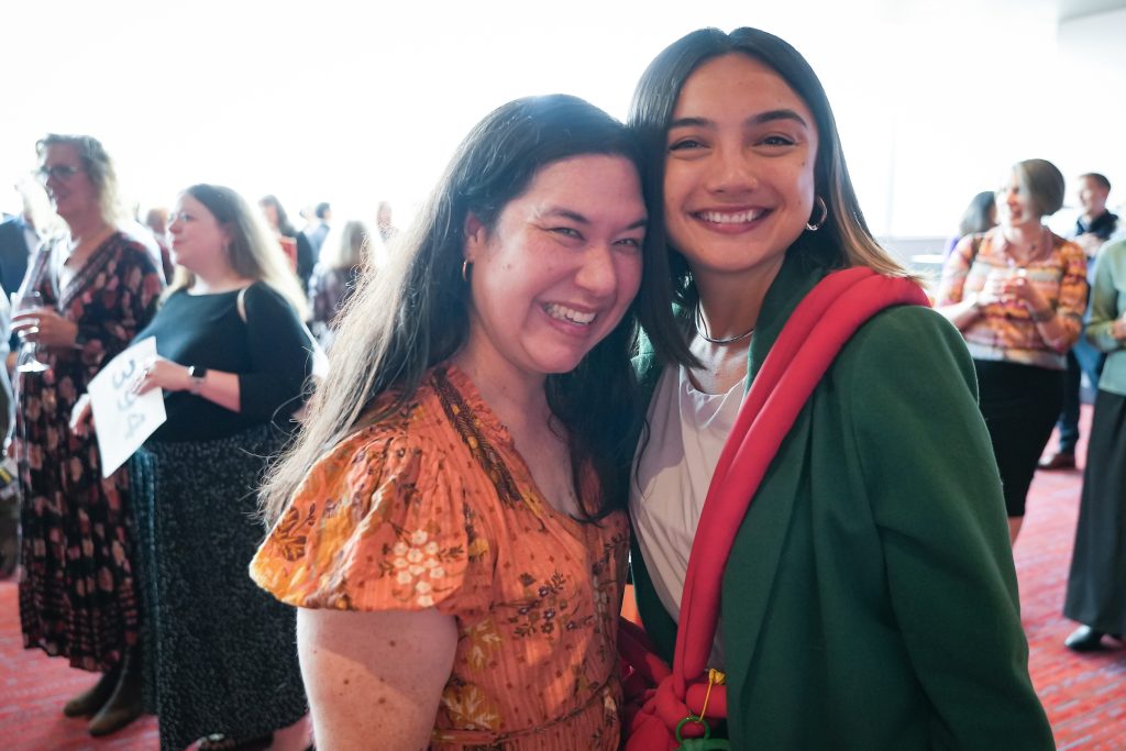 Two women posing closely together at ArtsFund's 2025 Celebration of the Arts with other attendees mingling in the background.